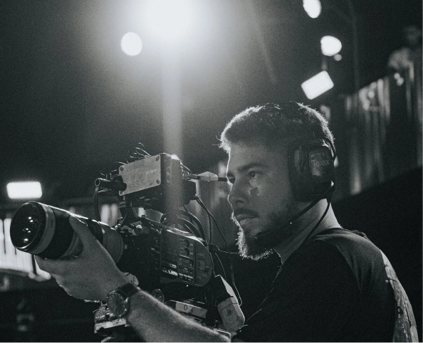 Man with beard wearing headphones operating a professional video camera in a dimly lit setting.