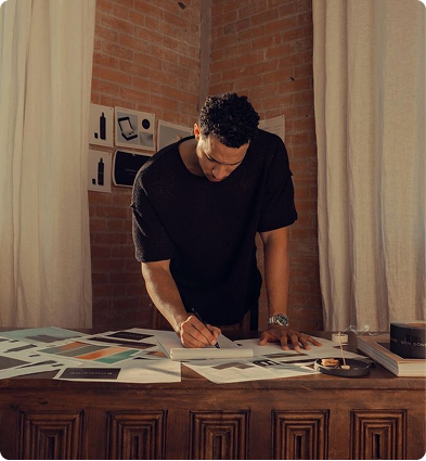 Man leaning over desk sketching or writing on paper in a room with brick walls and curtains.