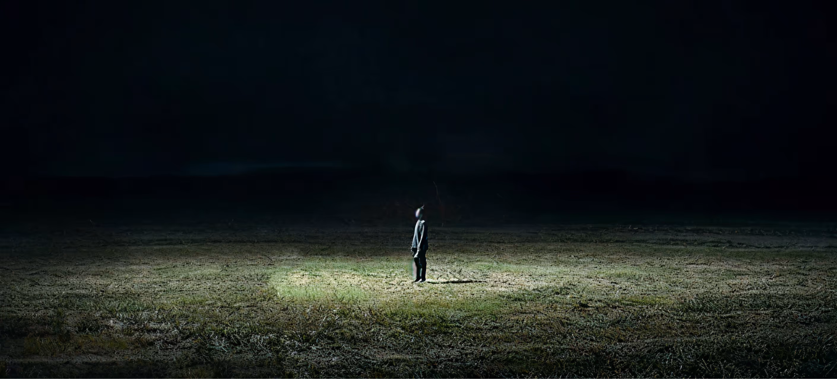 Person stands alone under light in a dark, open grassy field at night.