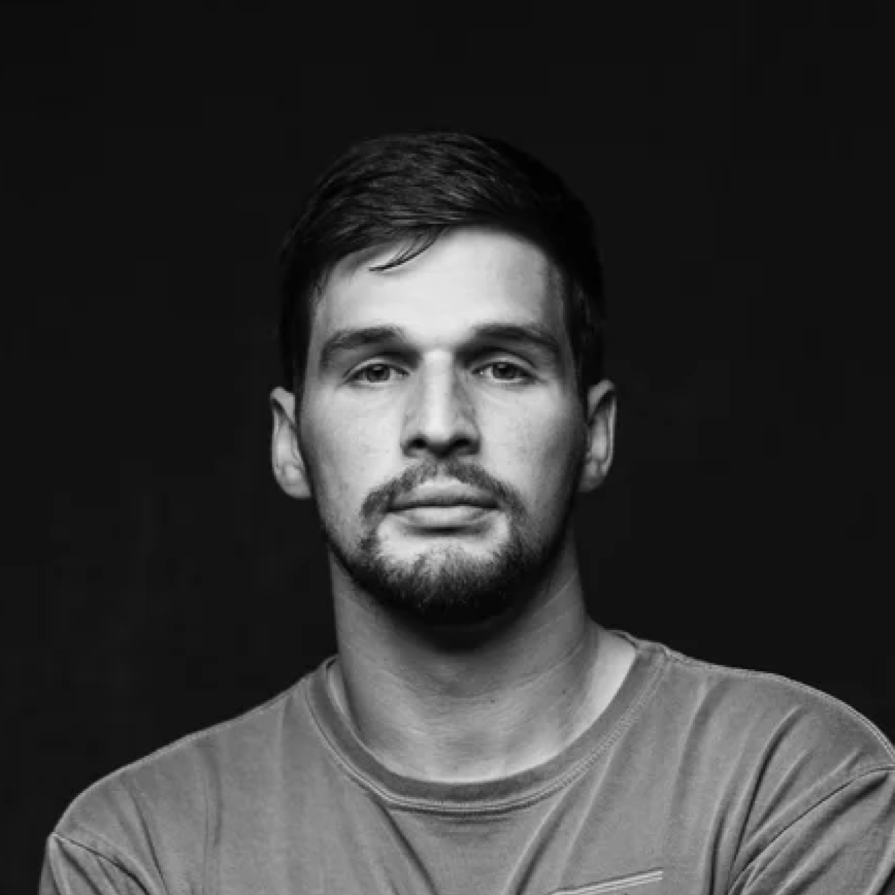 Black and white portrait of a serious man with short dark hair and a beard wearing a plain t-shirt.