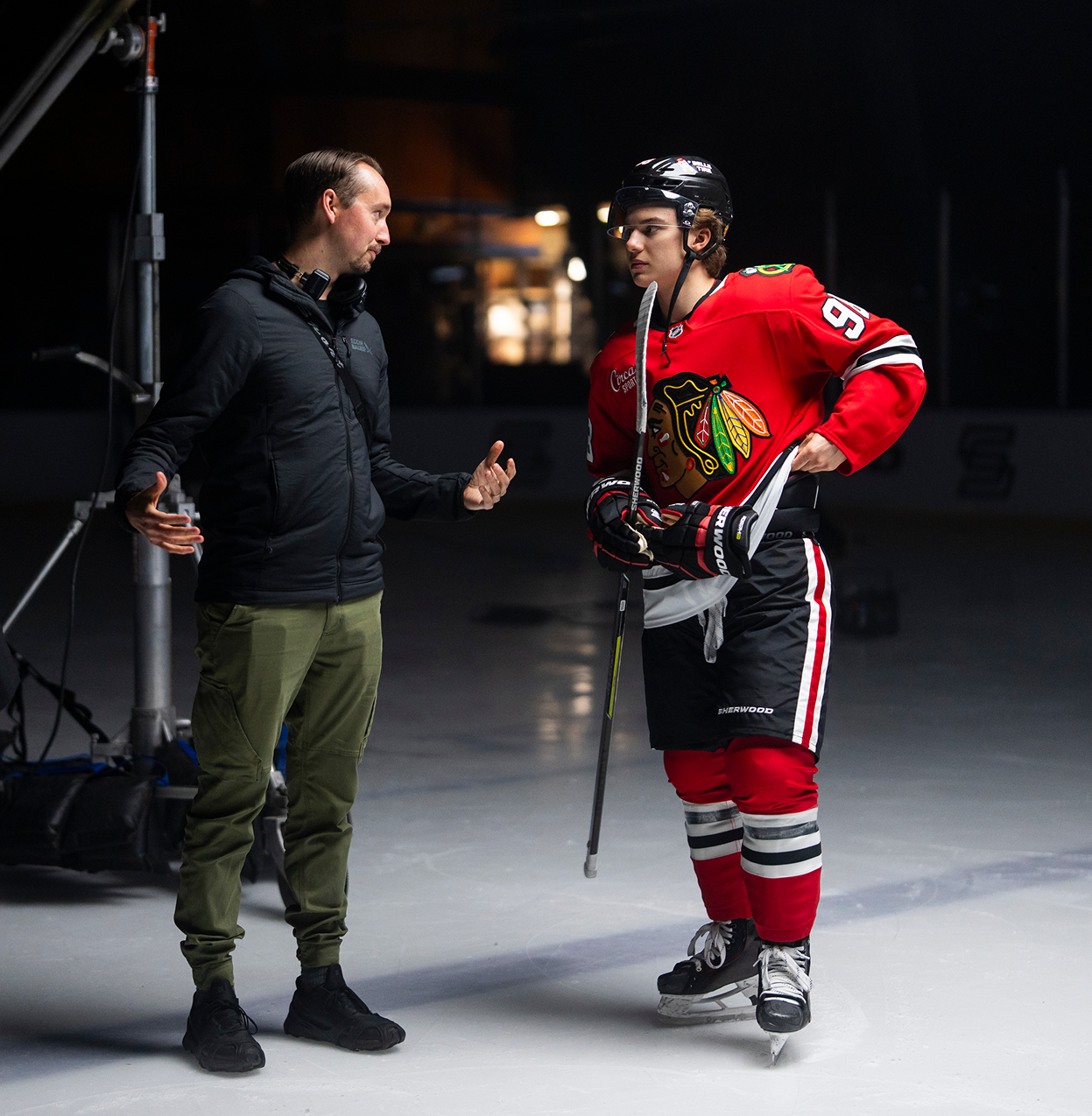 Man in black jacket talks to ice hockey player in red Chicago Blackhawks jersey on ice rink.