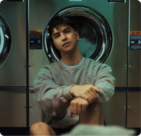 Young man in gray sweater sitting in front of washing machines in a laundromat.