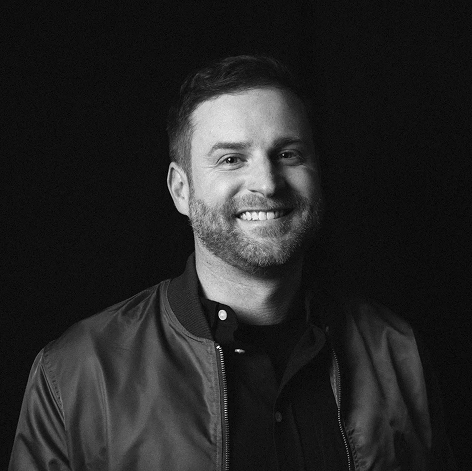 Smiling man with short hair and beard wearing a zip-up jacket and buttoned shirt, black background.