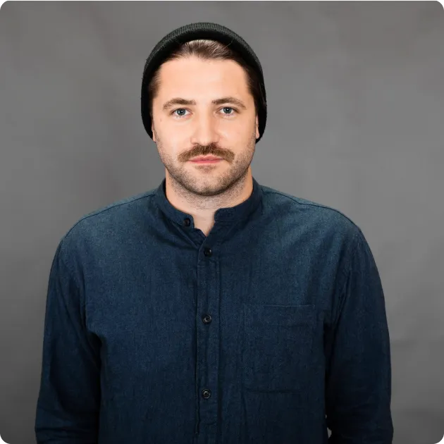 Portrait of a man with a mustache wearing a black beanie and a dark blue button-up shirt against a gray background.