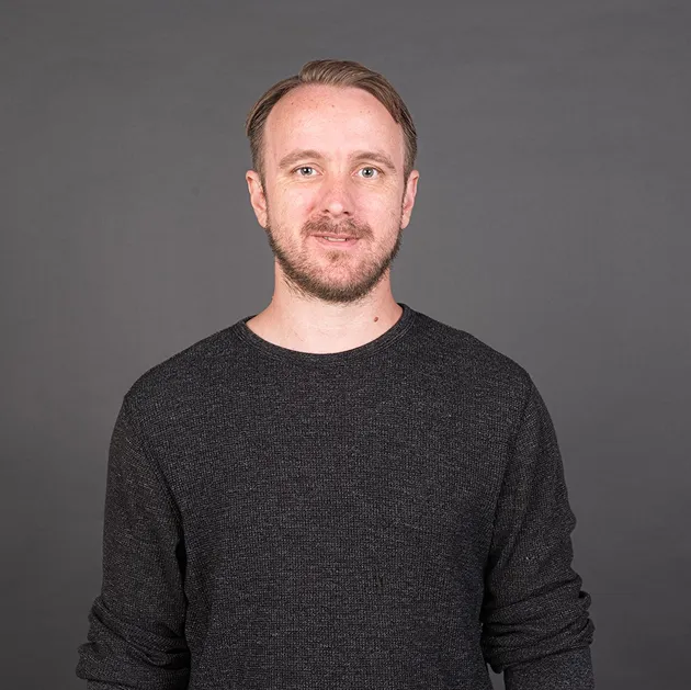 Man with short brown hair and beard wearing a dark gray sweater against a gray background.