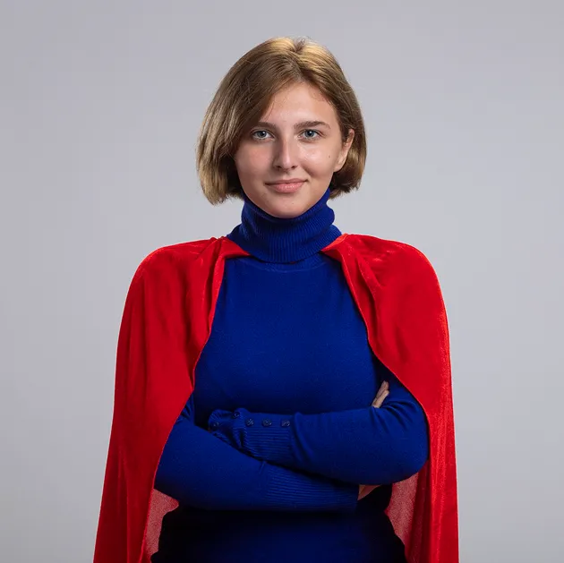 Young woman with short blond hair wearing a blue turtleneck sweater and a red cape, standing with arms crossed against a plain background.