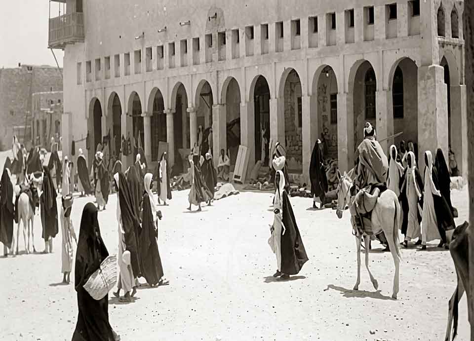 مجموعة من الناس يرتدون ملابس تقليدية ويسيرون أمام مبنى بأقواس حجرية في منطقة صحراوية.