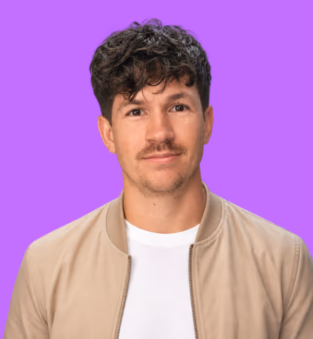 Ein Mann mit kurzen braunen Haaren, einem Bart und beigen Jacke vor lilanem Hintergrund