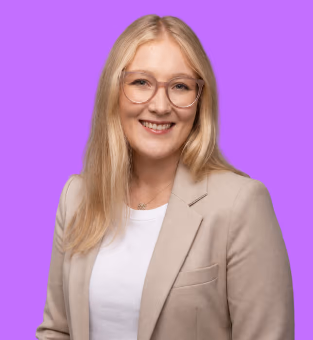 Eine junge Frau mit langen blonden Haaren, einer Brille und einem beigen Blazer vor einem lilanen Hintergrund.