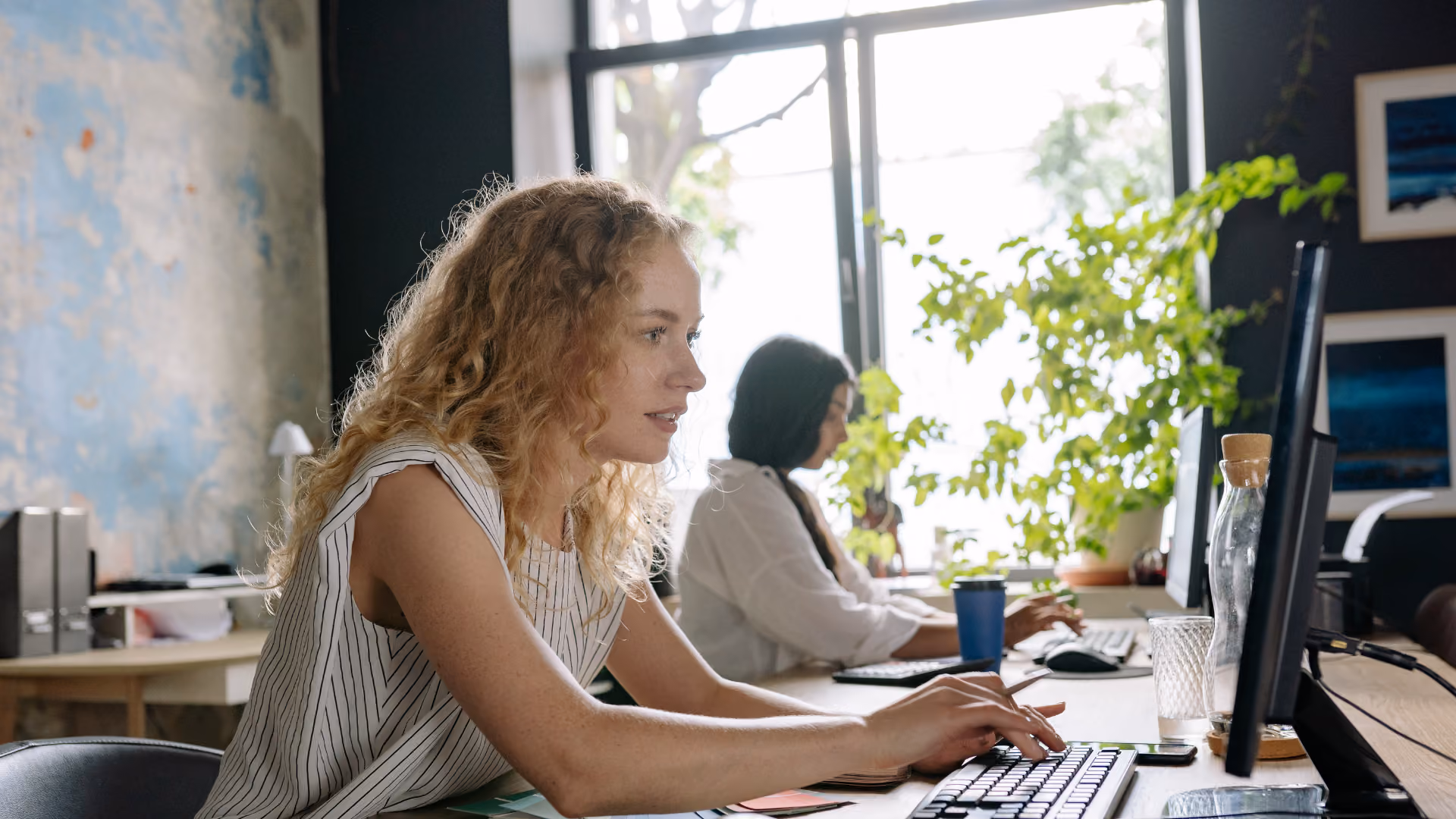 Zwei Frauen sitzen an Schreibtischen und arbeiten an Computern in einem hellen Büro mit Pflanzen im Hintergrund