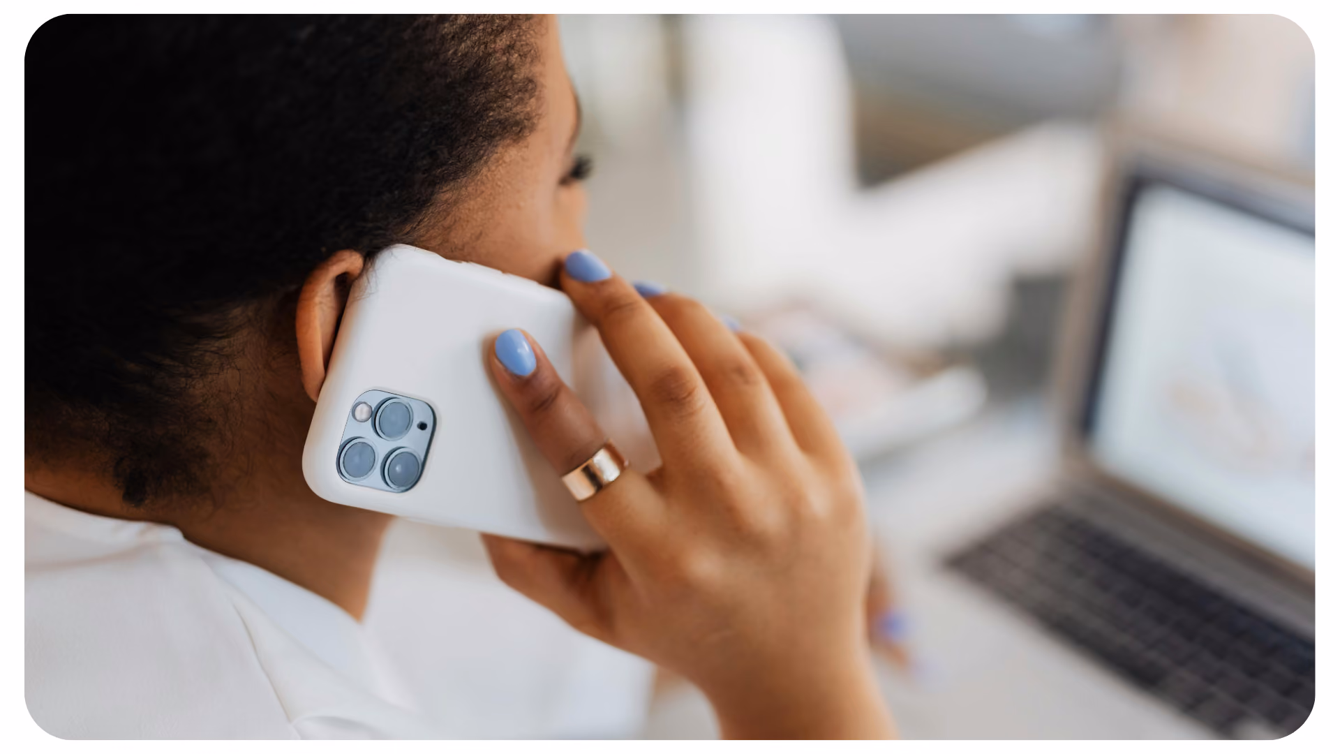 Person mit blauer Nagellackierung hält ein weißes Smartphone ans Ohr und blickt auf einen Laptopbildschirm
