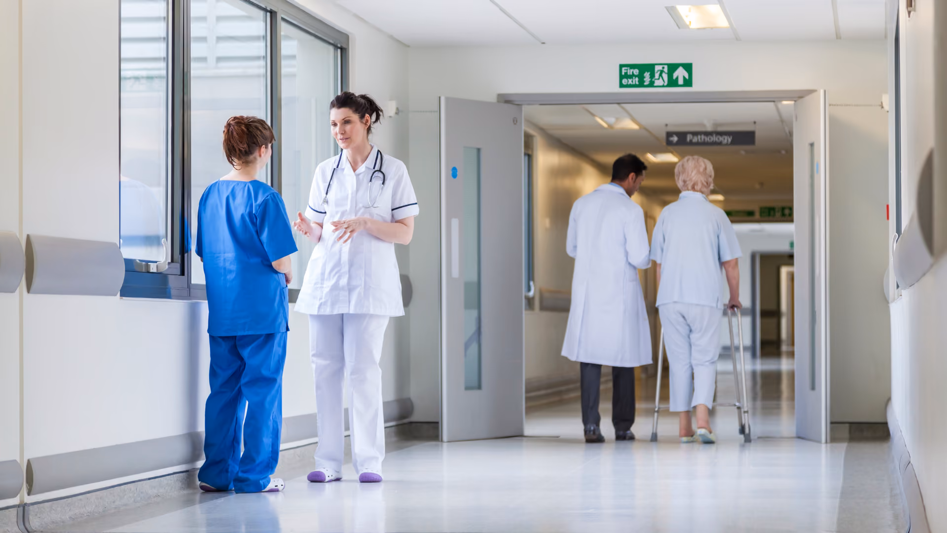 In einem Flur einer Klinik unterhalten sich eine Ärztin und MFA. Im Hintergrund begleitet ein Arzt eine Patientin mit Rollator.