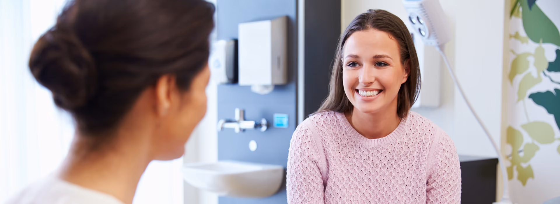Ärztin sitzt einer Patientin im rosa Oberteil gegenüber, die lächelt.
