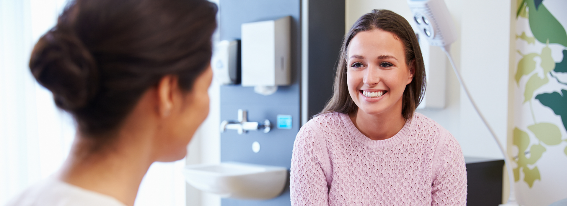 Ärztin sitzt einer Patientin im rosa Oberteil gegenüber, die lächelt.
