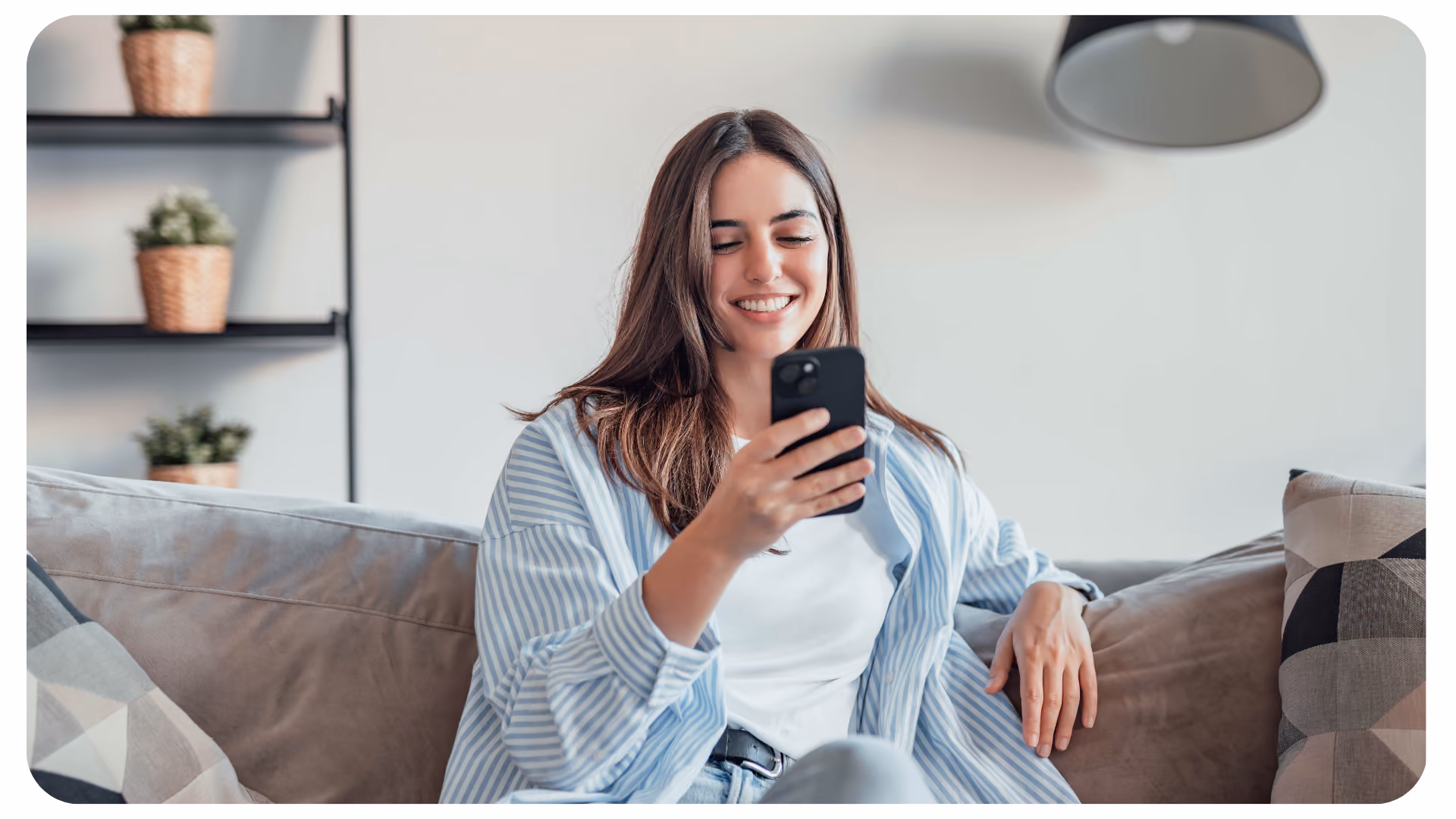 Frau mit weißem Shirt und blau gestreifter Bluse sitzt auf der Couch und hält ein Handy in der Hand.