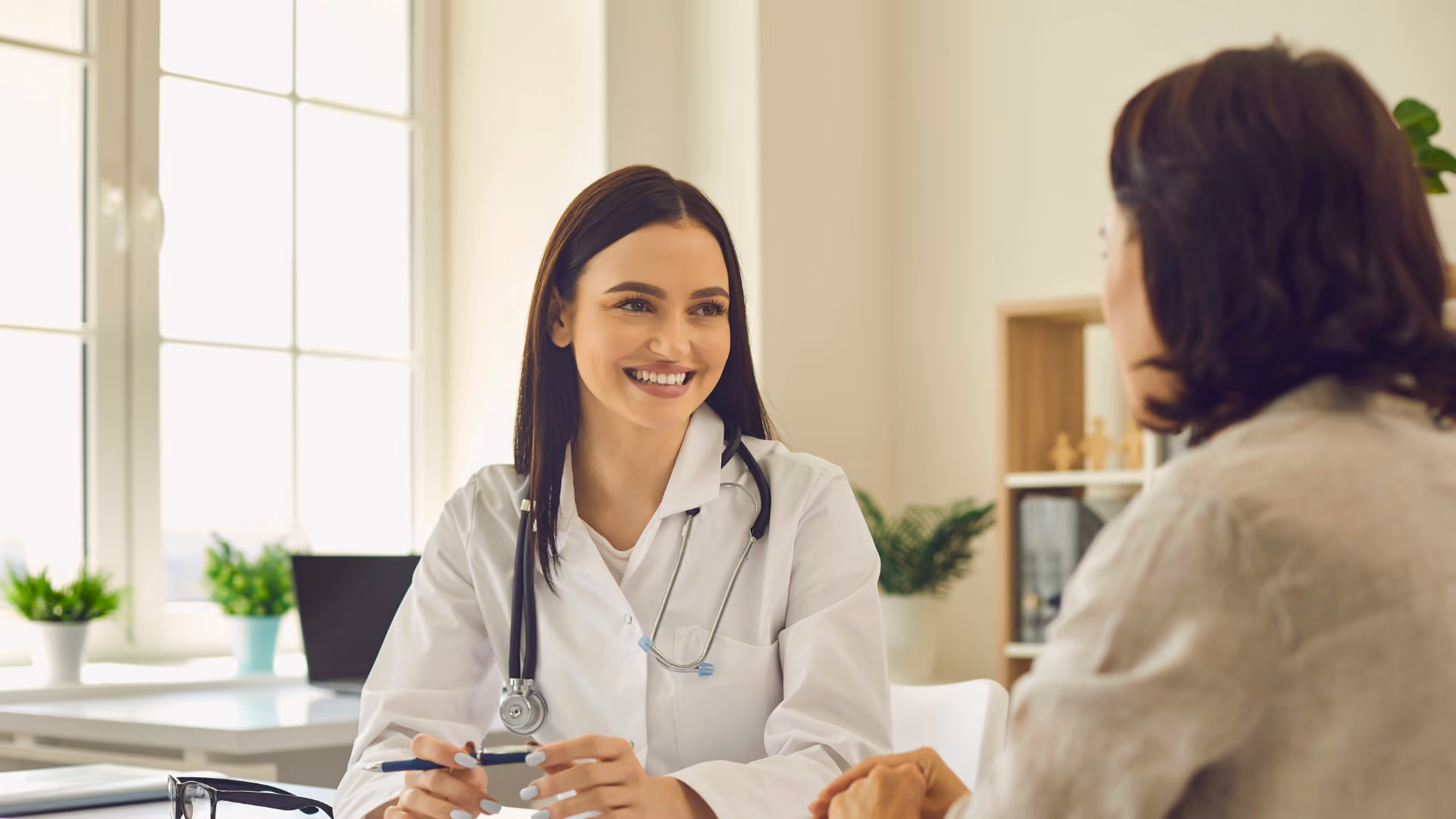 Eine Ärztin mit langen dunklen Haaren sitzt einer Patientin gegenüber und lächelt.