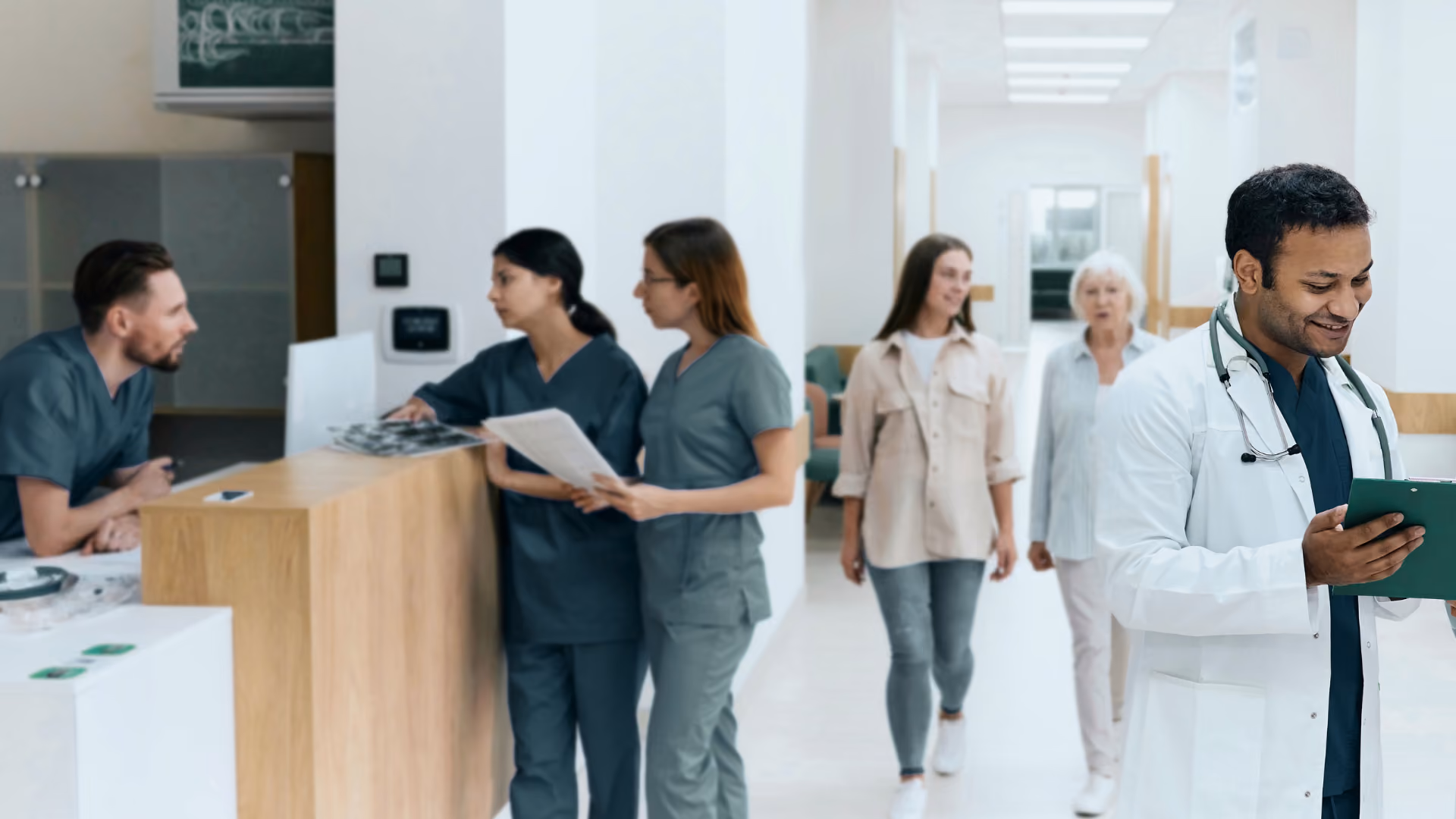 Mehrere medizinische Fachkräfte im Gespräch an einem Empfangstresen in einem hellen Krankenhausflur, im Vordergrund ein Arzt mit Tablet.