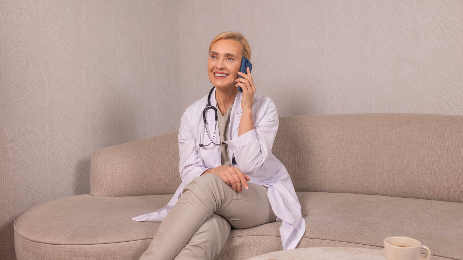 Eine Ärztin sitzt auf einer beigen Couch und telefoniert.