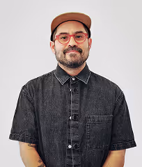 Man with beard, red glasses, and brown cap wearing a black button-up shirt against a plain background.