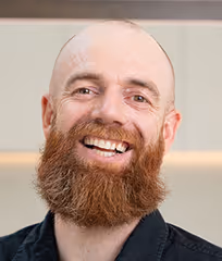 Smiling bald man with a full red beard wearing a dark shirt against a plain background.
