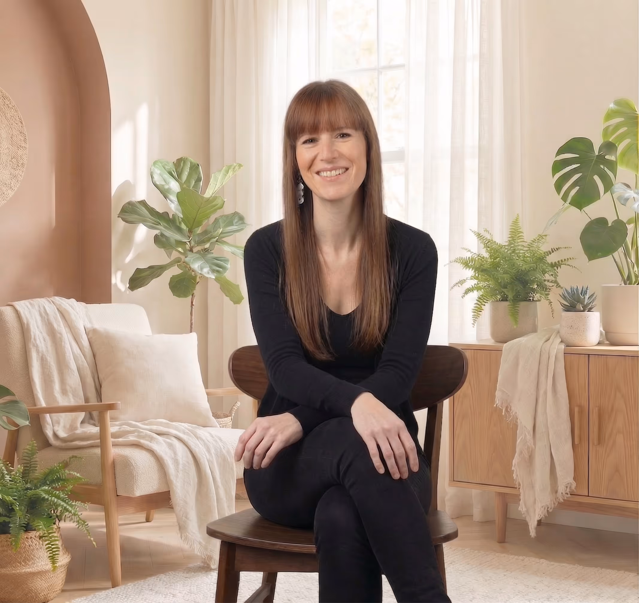 Lächende Frau mit langen braunen Haaren, die auf einem Stuhl in einem hellen, gemütlichen Raum mit Pflanzen sitzt.