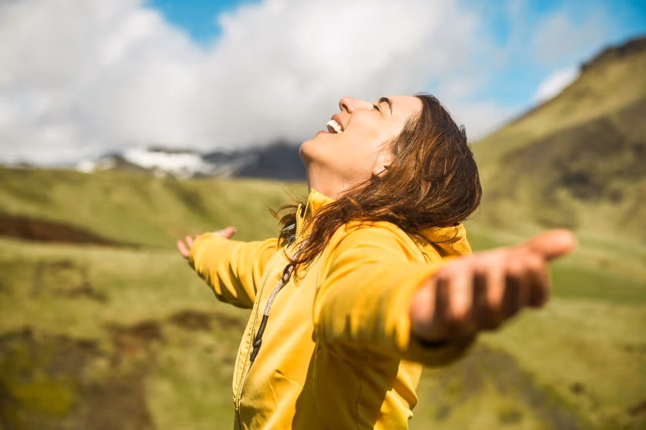 Frau in gelber Jacke genießt die Natur mit ausgebreiteten Armen in einer grünen Berglandschaft.