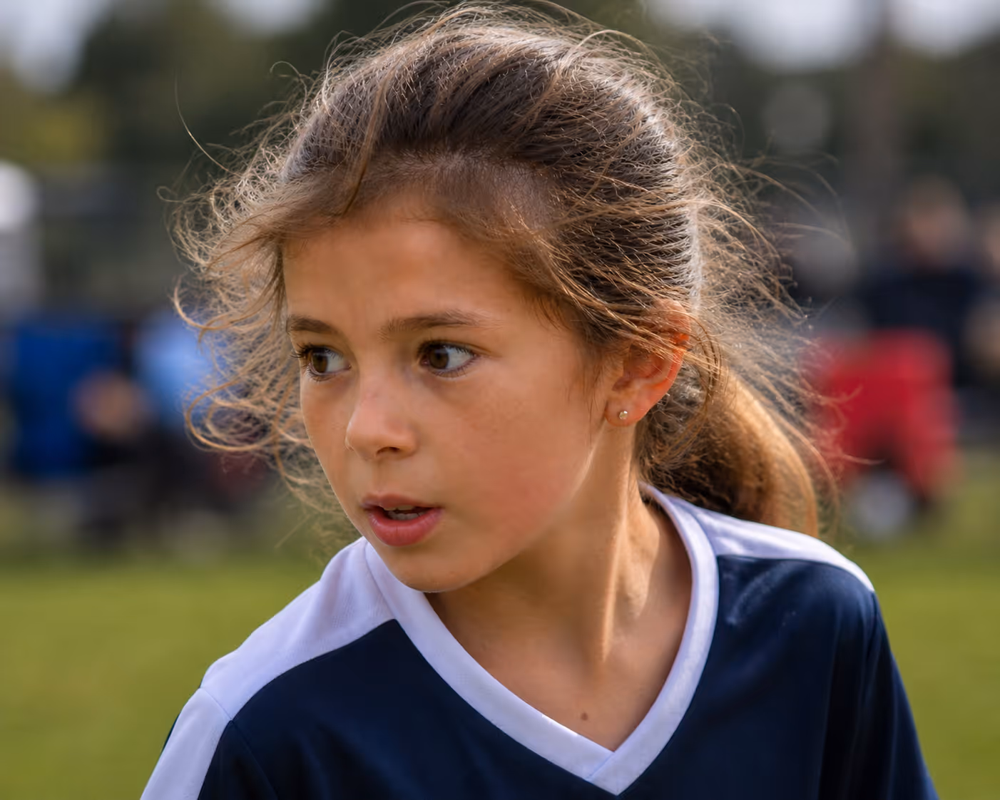 Young girl with brown hair in a ponytail wearing a navy and white sports jersey outdoors.