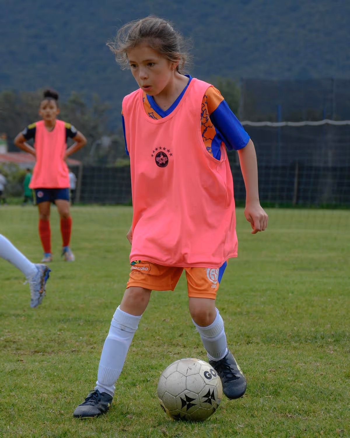 Young girl in orange shorts and pink training bib dribbling a soccer ball on a grassy field.
