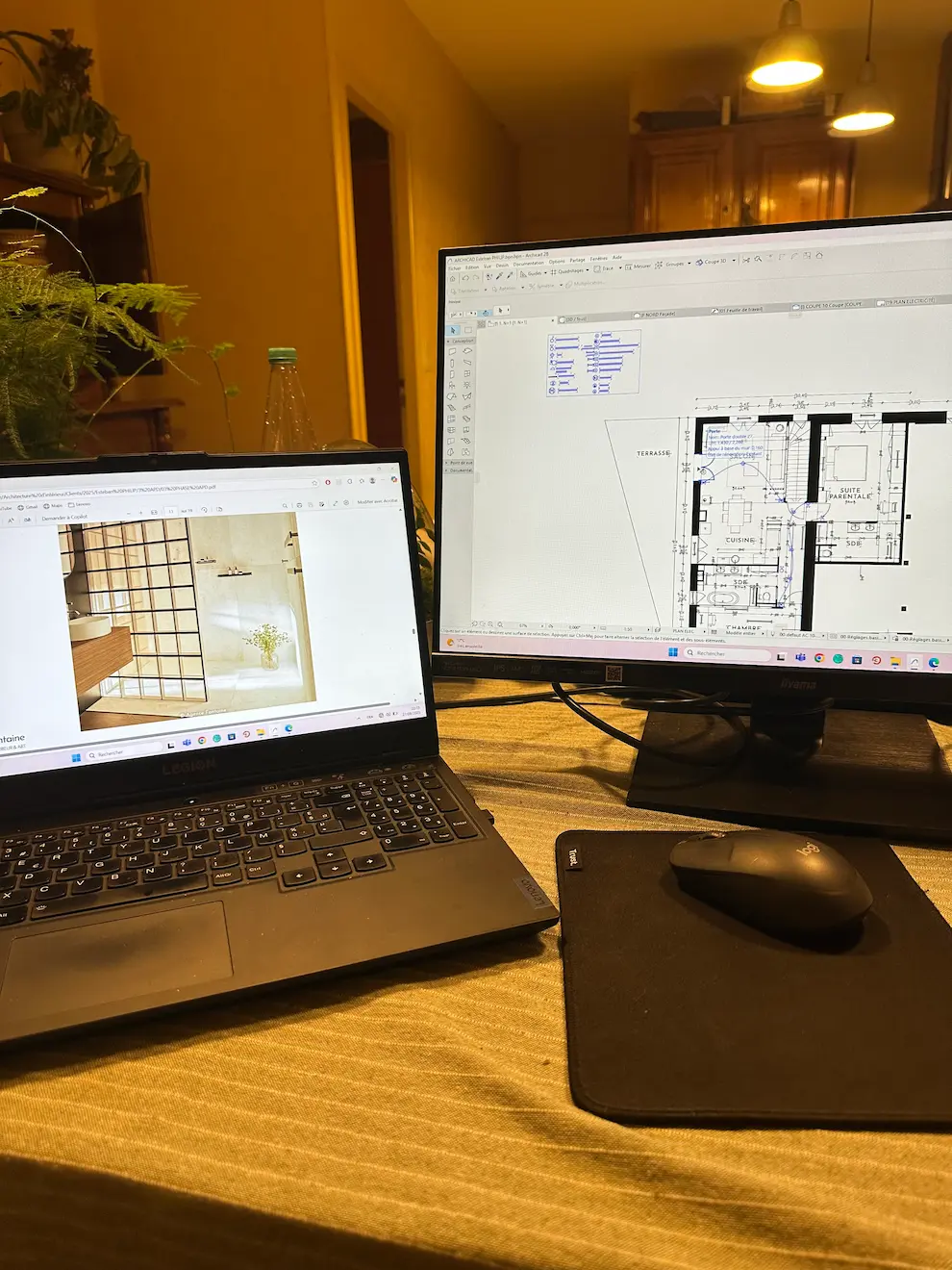 Desk with laptop showing bathroom image and monitor displaying architectural floor plan beside mouse on mat.