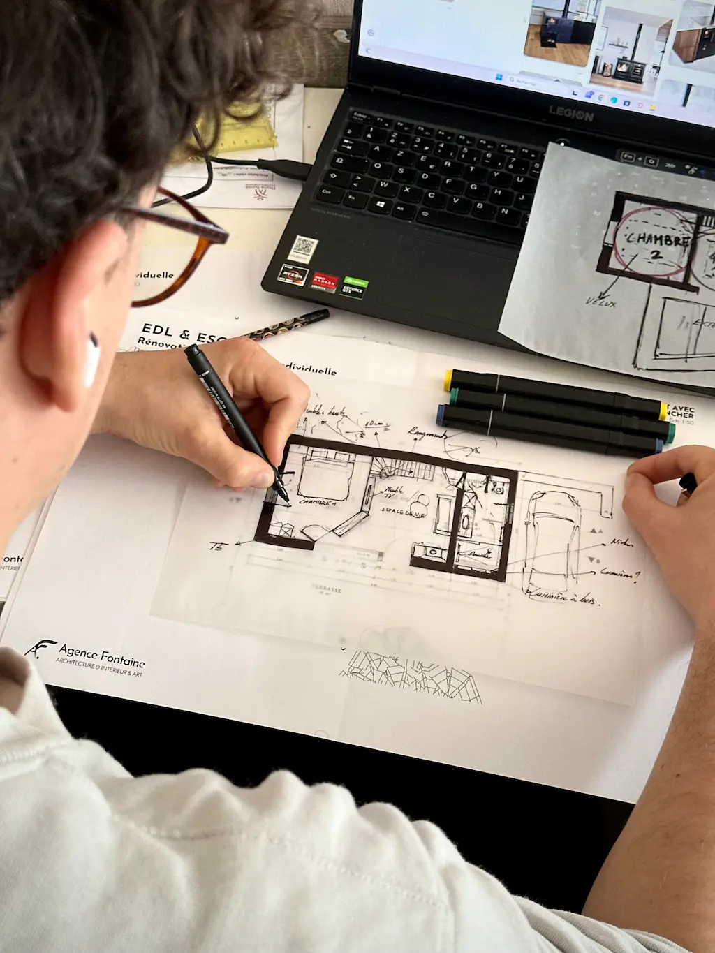 Architecte travaillant sur un plan de maison avec des marqueurs et un ordinateur portable.