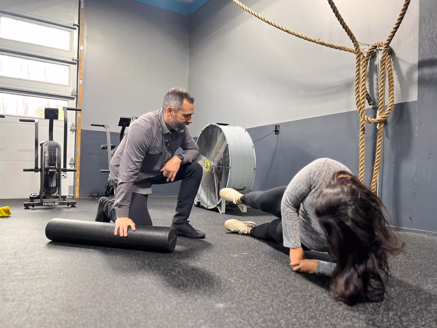 Man kneeling with foam roller watches woman in gray top doing side leg raises on gym floor.