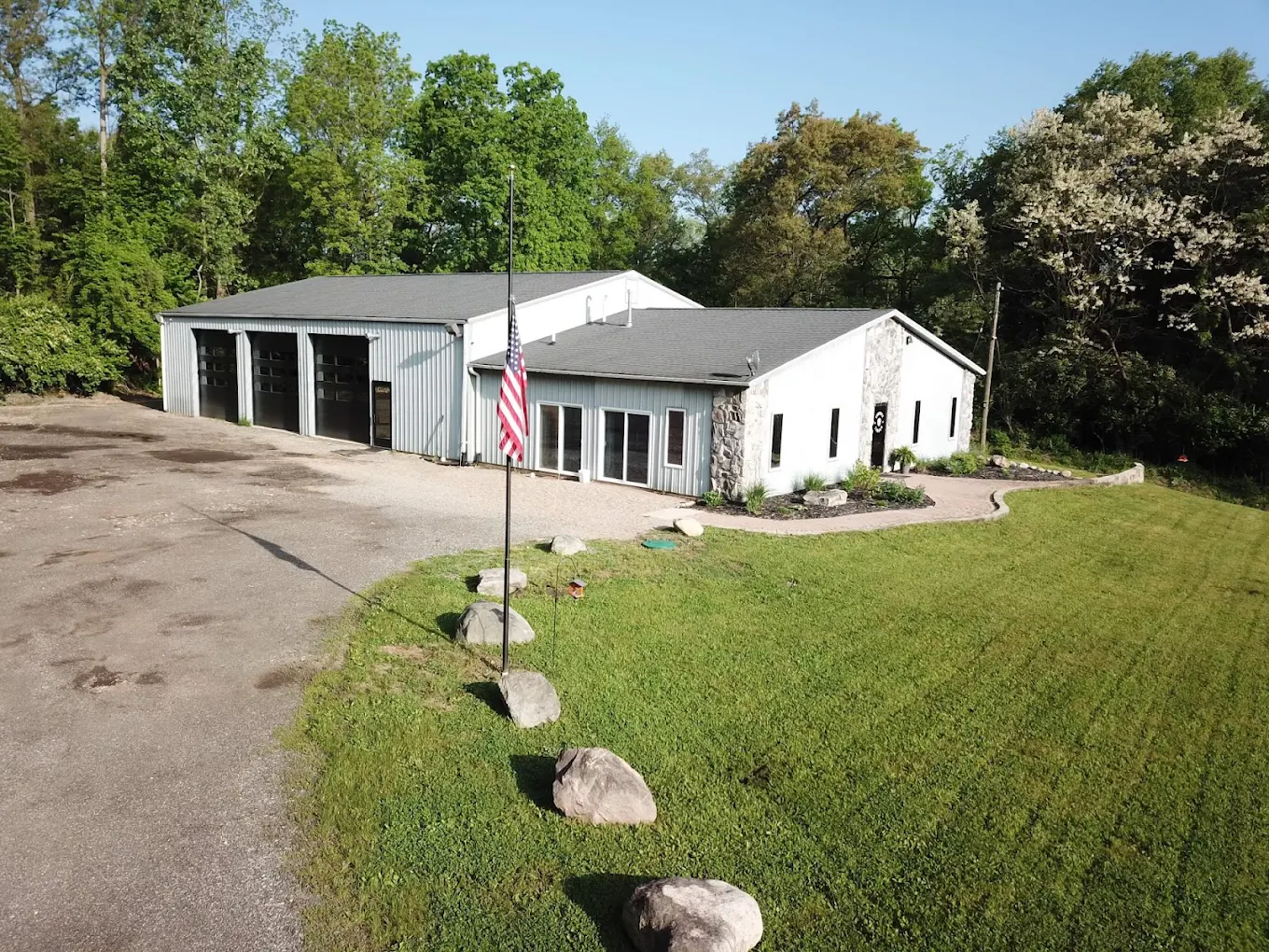 White commercial building with gray roof, garage bays, American flag, and green lawn surrounded by trees.