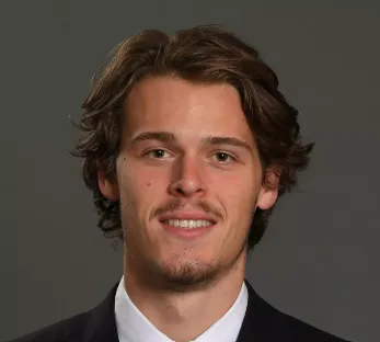 Young man with medium-length brown hair and light facial hair wearing a suit and white shirt.