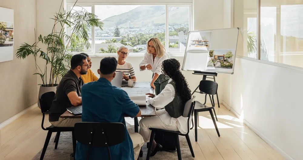 A group of five people having a meeting in a bright room with large windows and a presentation board.