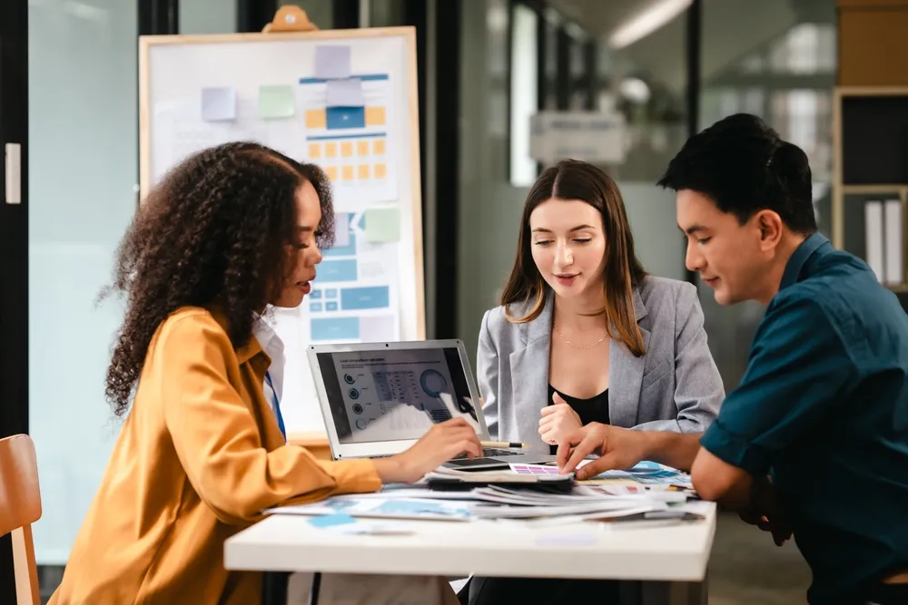 Three colleagues collaborate over documents and a laptop in a modern office meeting room.