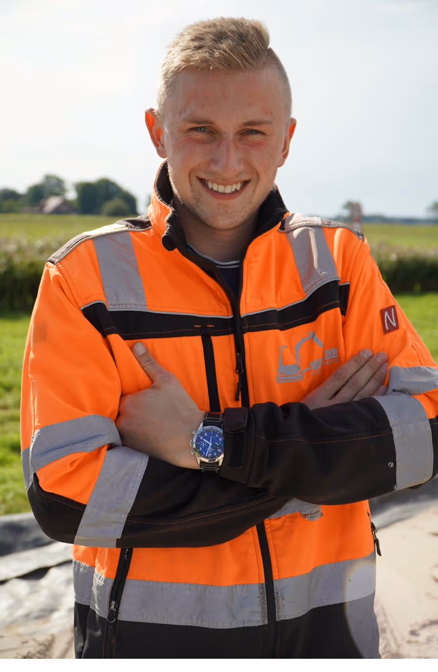 Junger Mann in orange-schwarzer Arbeitsjacke lächelt mit verschränkten Armen vor einer Wiese.