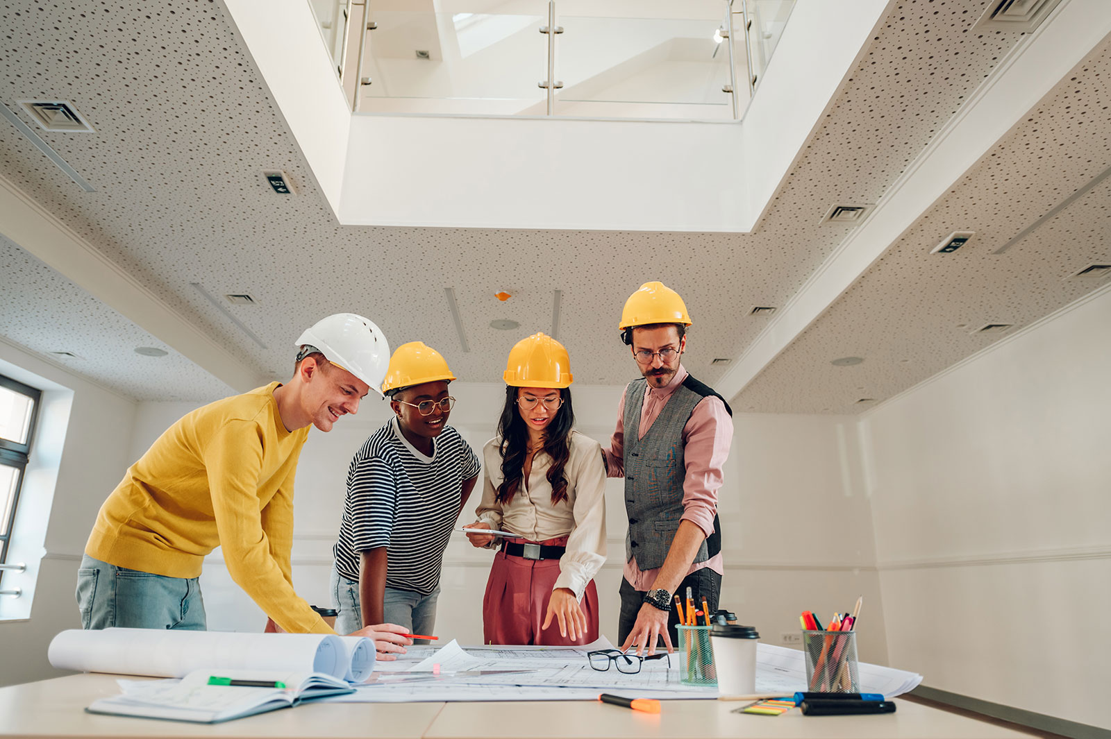 Four architects or engineers wearing hard hats discussing blueprints on a table in a bright, modern office.