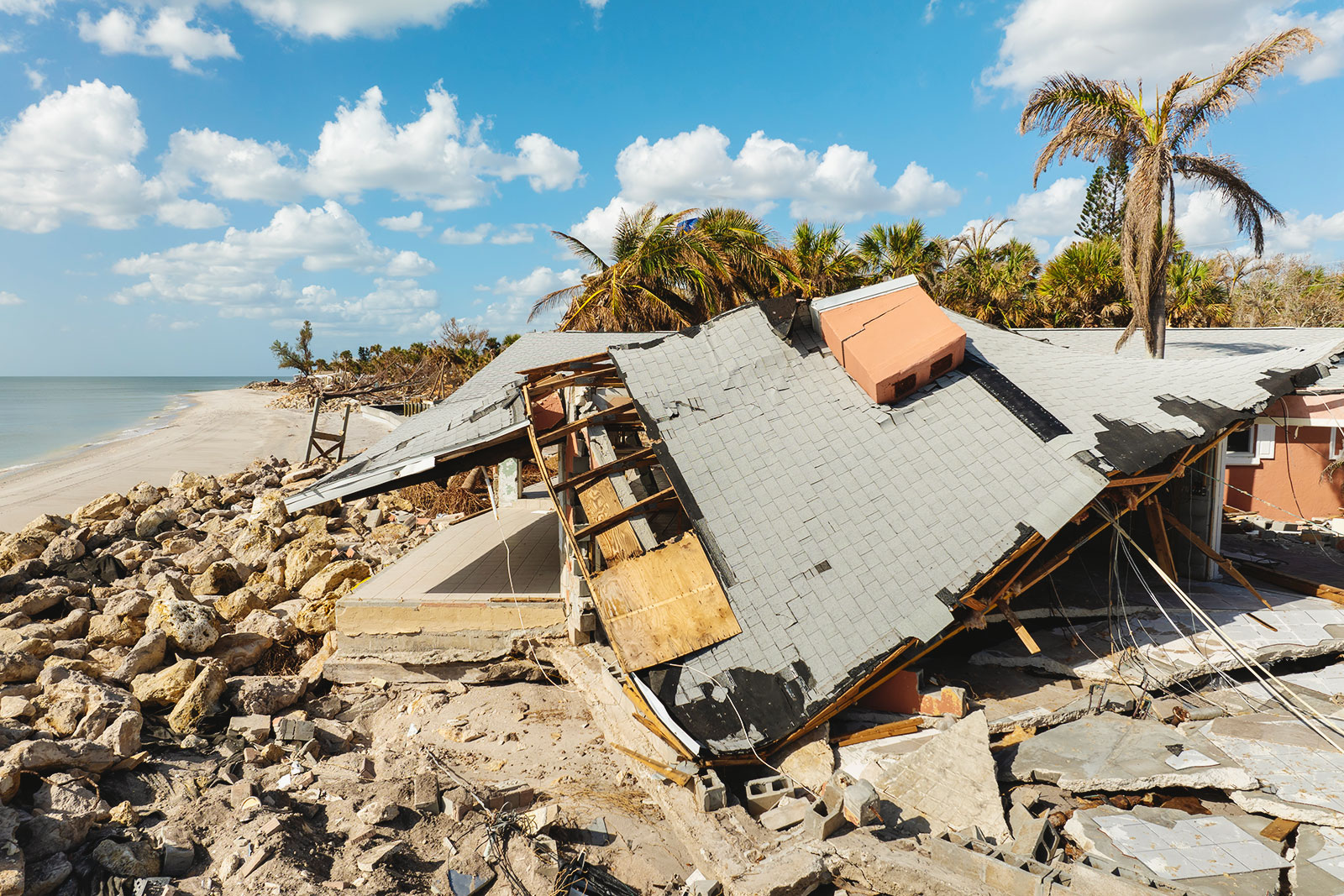 Collapsed house with roof damage and rubble near a rocky beach under a partly cloudy sky.