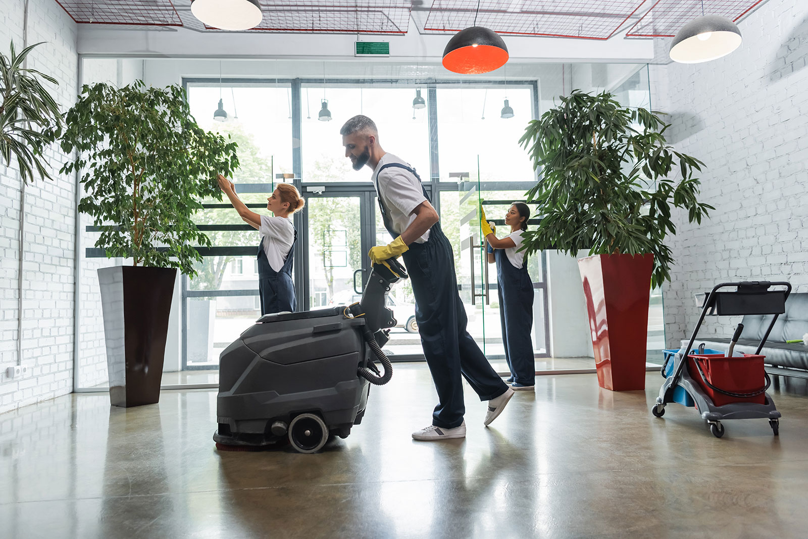 Three janitors cleaning a bright office lobby with floor scrubber, wiping glass doors, and dusting plants.