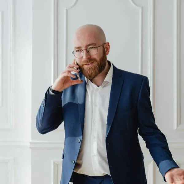 Homme chauve avec lunettes et barbe portant un costume bleu parlant au téléphone dans un bureau.