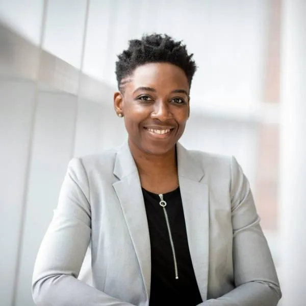 Femme souriante portant un blazer gris clair et un haut noir posant devant une fenêtre moderne.