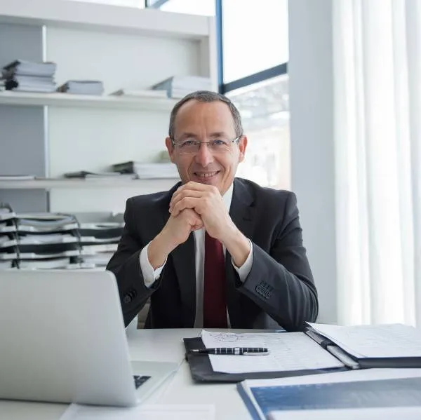 Homme d'affaires souriant assis à un bureau avec ordinateur portable et documents en arrière-plan.