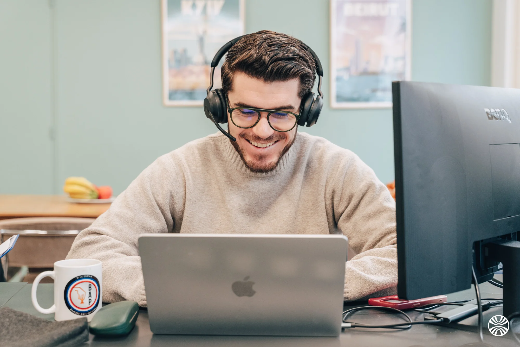 Homme souriant avec casque audio, lunettes, et pull, travaillant sur un ordinateur portable Apple.