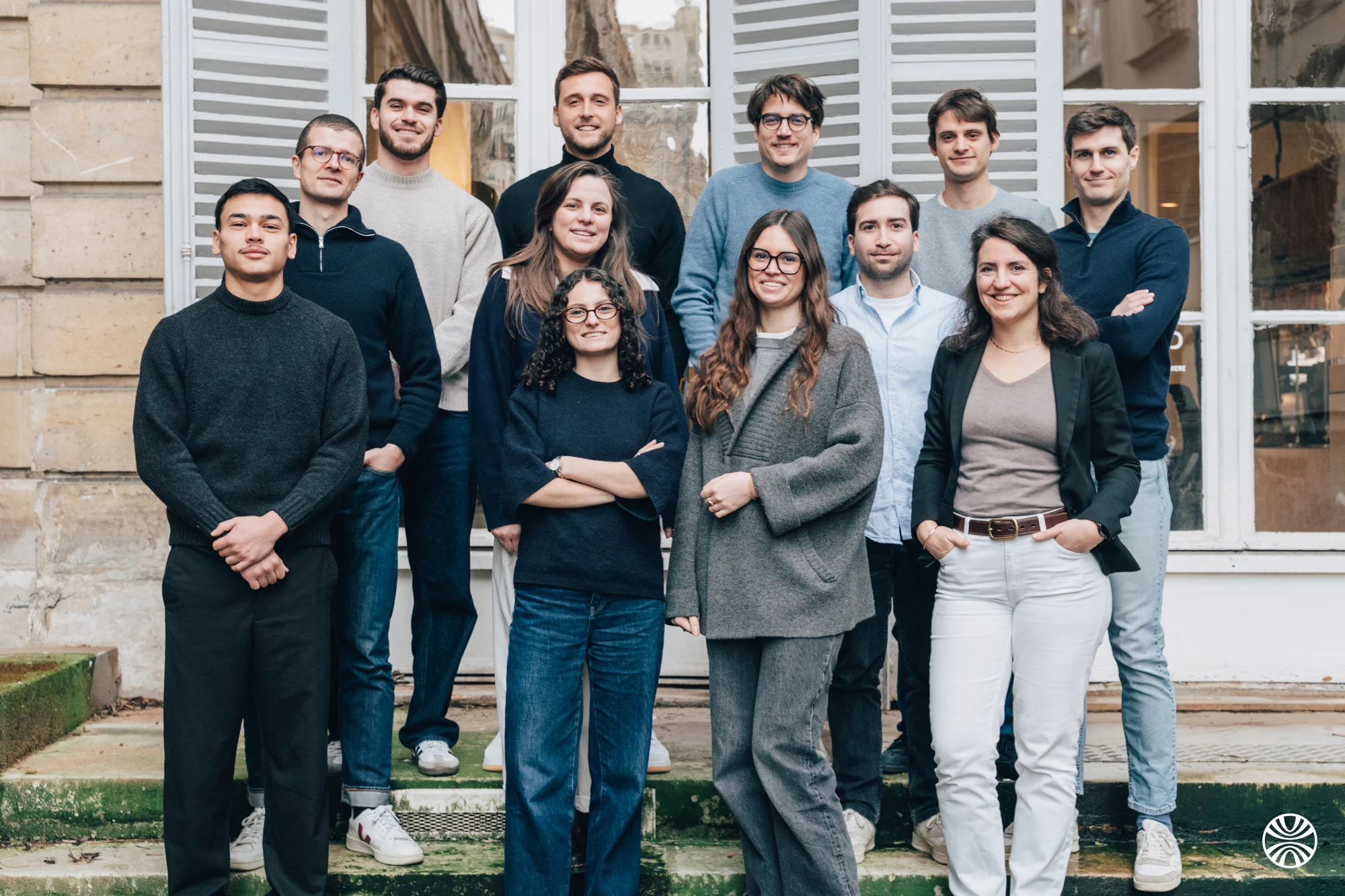 Groupe de 13 personnes souriantes posant devant un bâtiment avec des fenêtres blanches.