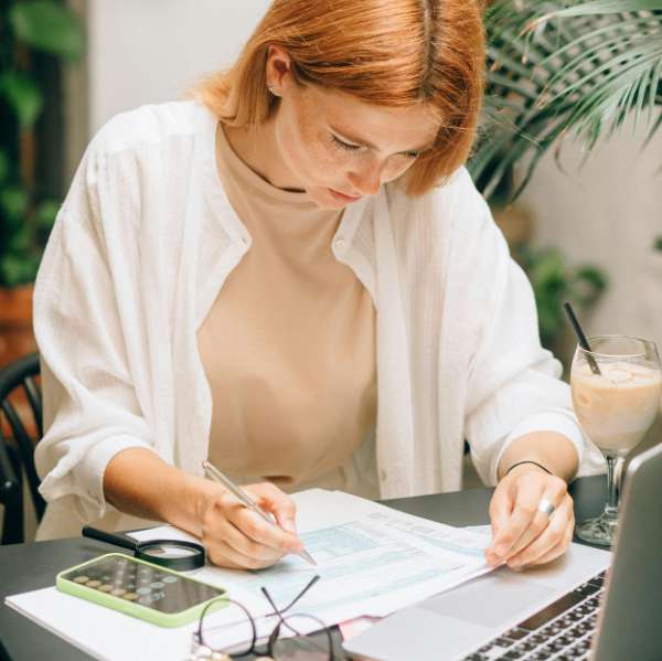 Femme aux cheveux roux concentrée écrivant sur un document avec une calculatrice et un ordinateur près d'elle.
