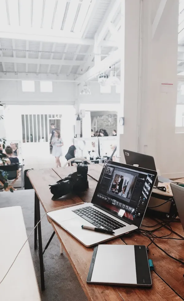 Wooden table with a laptop showing photo editing software, a camera, and a digital drawing tablet in a bright workspace.