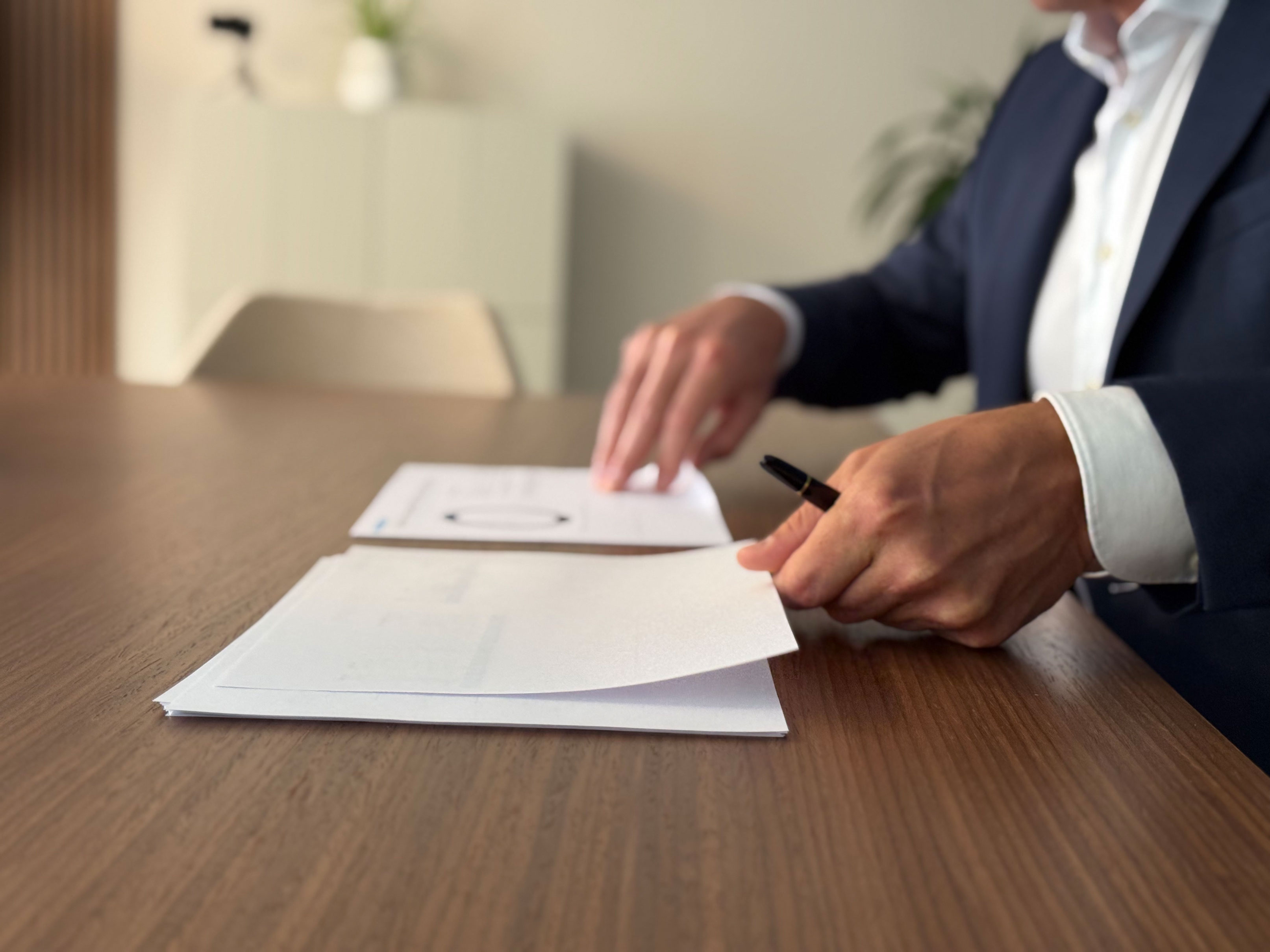 Person in business attire sitting at a wooden table, reviewing and handling paperwork with a pen in hand.