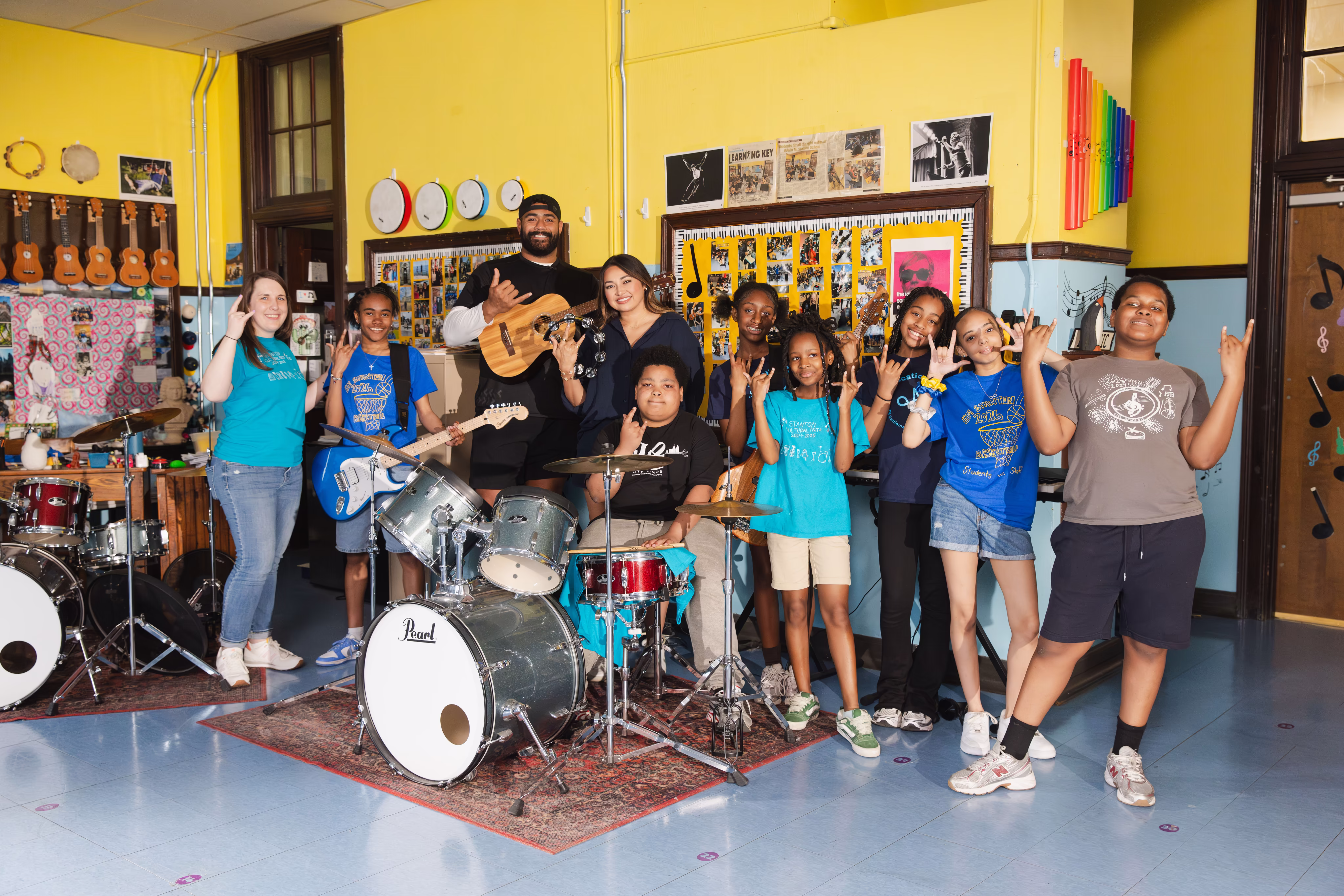 A diverse group of children and two adults posing with musical instruments like drums, guitars, and tambourine in a colorful music classroom.