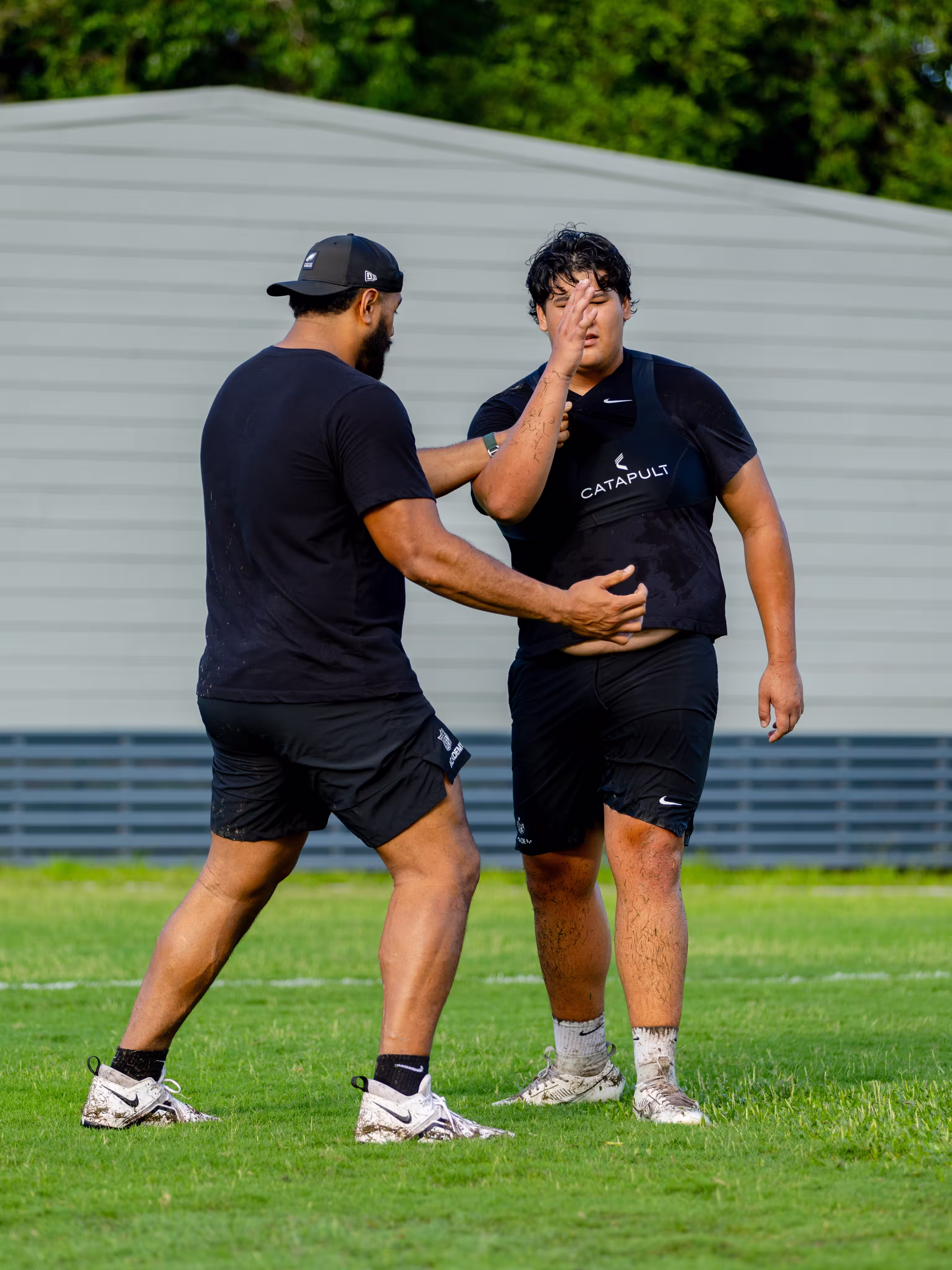 Two men in black athletic wear and muddy white Nike cleats engage in a training drill on a grassy field, one gesturing with his hands.