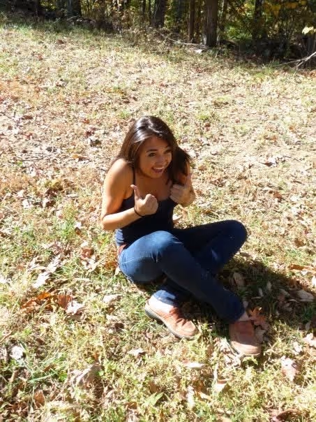 Young woman sitting on grass outdoors, giving two thumbs up and smiling.